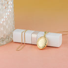 oval gold locket with border engraving leaning against a white lipstick container on a peach and pink background