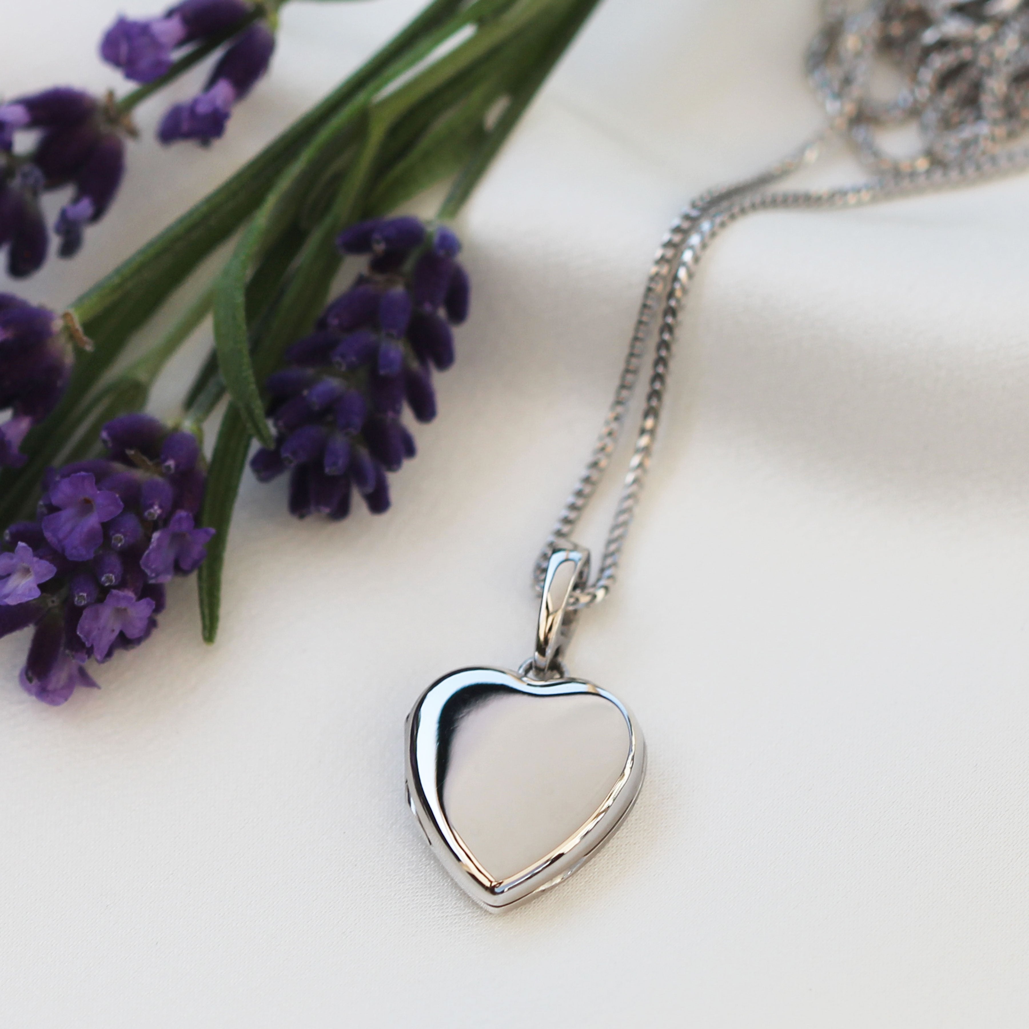 An 18 ct white gold heart locket lying alongside a spray of purple flowers, on a bed of white silk.